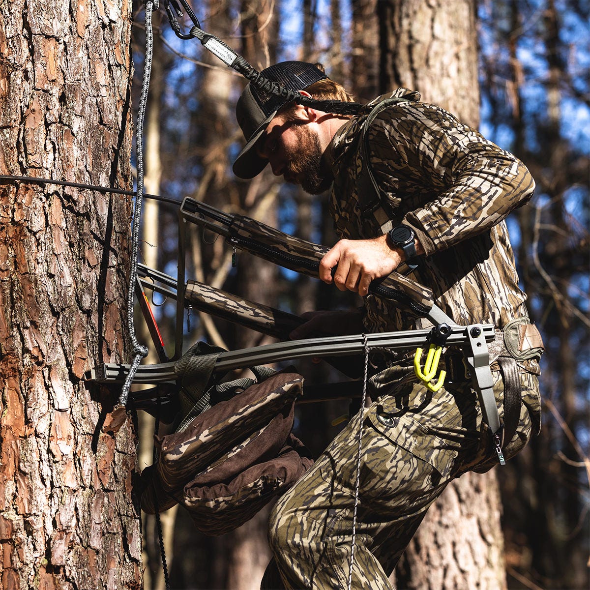 Man in camo ascends a tree using the Summit Dual Threat PRO SD climbing treestand. He is gripping the side arms and has his weight on the adjustable front bar.