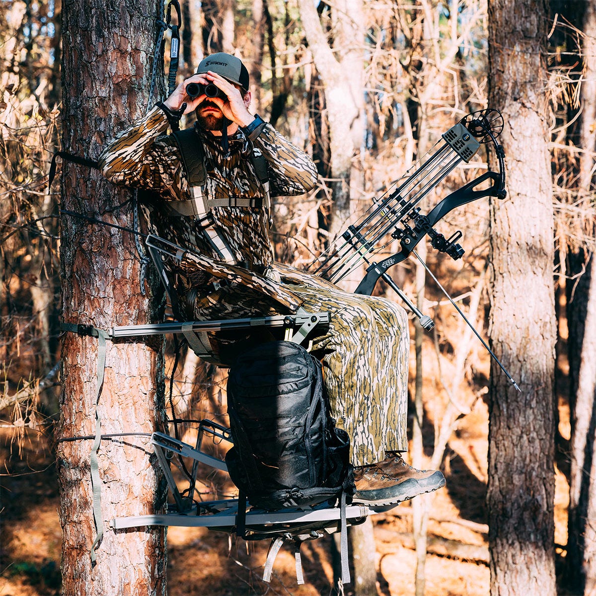 View of a man sitting in the Summit Dual Threat PRO SD climbing treestand in the Bow Hunting Position. He is looking sideways through his binoculars.