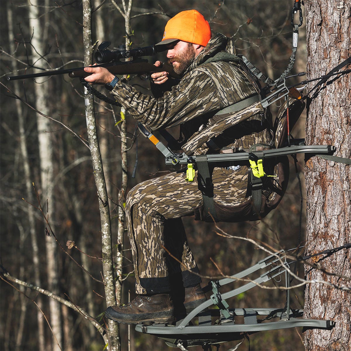 Side view of a man sitting in the Summit Dual Threat PRO SD climbing treestand in the Rifle Hunting position. He is using the front bar as a gun rest and pointing his rifle toward a target.